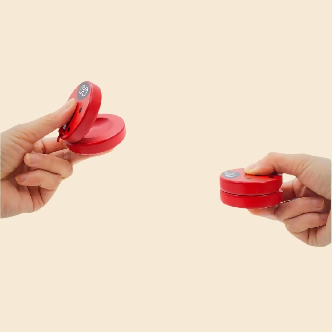 Two hands holding red ladybug castanets with a beige background. Demonstrating how to tap the castanet instruments.