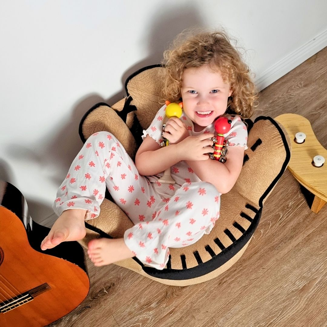 Child sitting on a toy chair holding two animal bell sticks, with a guitar on the floor.